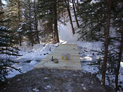 A completed timber bridge deck spans an icy creek in a snowy conifer forest, backlit by low sun.