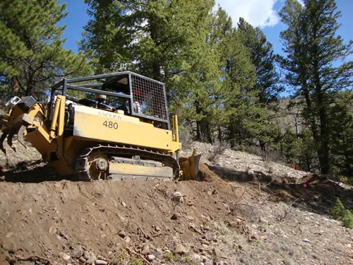 A yellow tracked trail dozer sits on freshly cut tread above a raw earth slope, silhouetted against a blue sky with scattered clouds.
