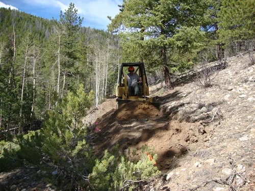 A small yellow trail dozer cuts fresh tread on a steep hillside surrounded by conifers and aspens, with a forested ridge behind.