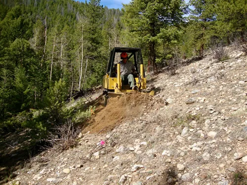 A small yellow tracked trail dozer cuts a fresh trail bench across a steep rocky slope, with a forested hillside rising behind.
