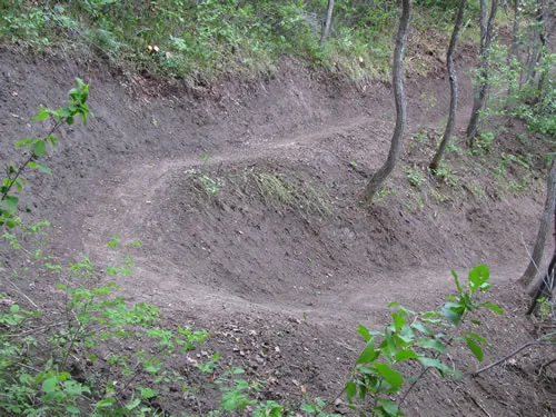 A freshly shaped bermed turn is dug into a wooded hillside, the banked outer wall clearly formed in raw soil.