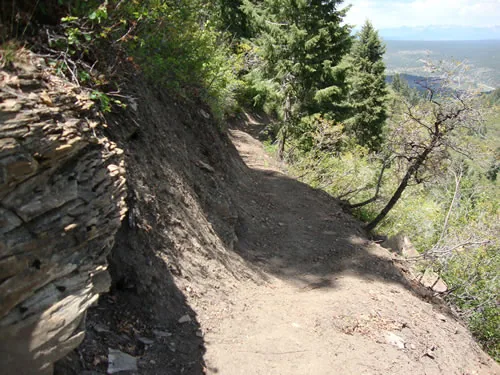 Dirt trail cut alongside a layered shale rock ledge, curving away downhill with a hazy valley beyond