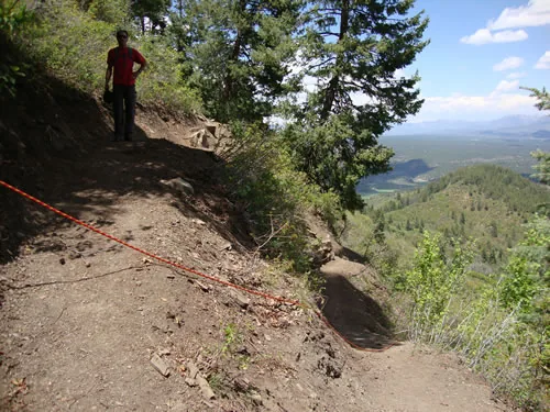 Person in a red shirt standing on a newly cut bench trail with an orange flag line strung along the alignment above a wide valley