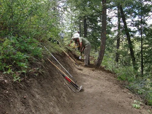 Trail worker shaping fresh tread on a hillside bench cut, with hand tools leaning against the uphill bank in the foreground