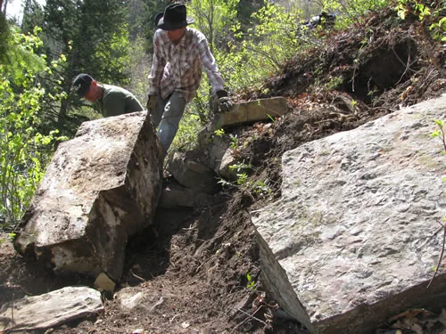 Two workers in wide-brim hats levering large stone slabs into place on a hillside