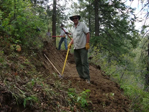 Trail worker in a wide-brim hat standing on a freshly cut bench holding a long-handled hand tool, with crew members further up the trail