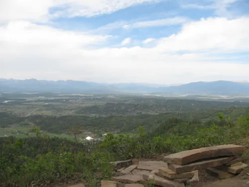 Wide view over a valley toward a distant mountain range under a cloud-streaked sky, with a flagstone overlook seat in the foreground