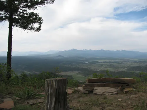 Panoramic view across a valley toward a distant mountain range, with a round flagstone seat and a cut stump in the foreground