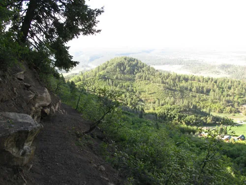Dirt trail cut across a steep slope past a pale rock outcrop, overlooking a forested ridge and hazy valley