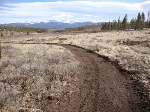 A newly cut singletrack curves through open sagebrush toward a distant snow-covered mountain range under scattered clouds.