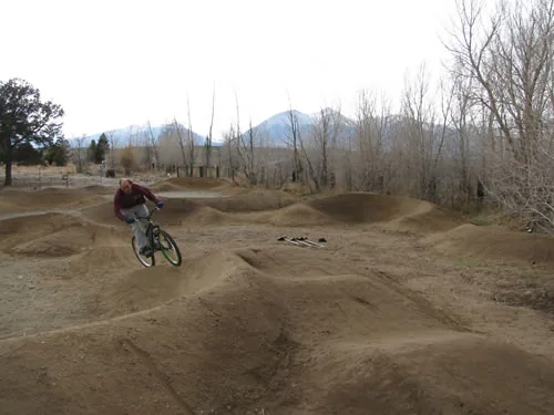 A mountain biker carves through the rollers of a dirt pump track with a mountain ridge in the distance.