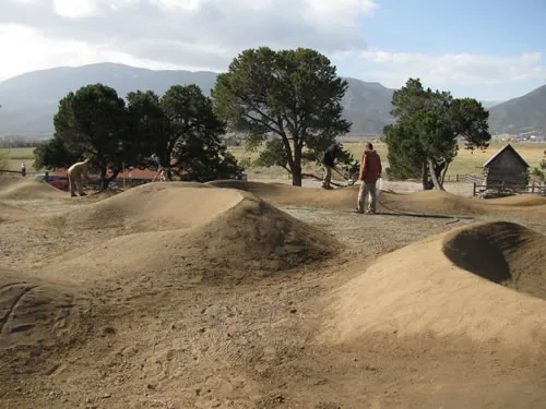 A dirt pump track of rolling humps and banked turns set among pinyon and juniper trees, with a rail fence and mountains behind.