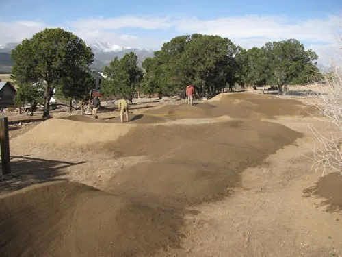 Sculpted dirt pump track rollers and banked turns in morning light, with people working in the background and snow-capped peaks on the horizon.