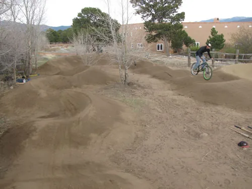 A rider lifts off a lip on a dirt pump track, with rolling berms in the foreground and a low building and mountains behind.