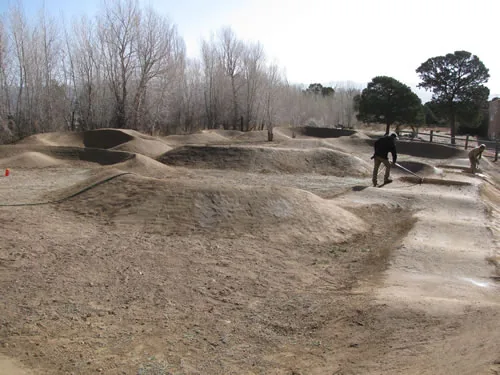 A worker rakes and shapes dirt rollers on a pump track under bare cottonwood trees.
