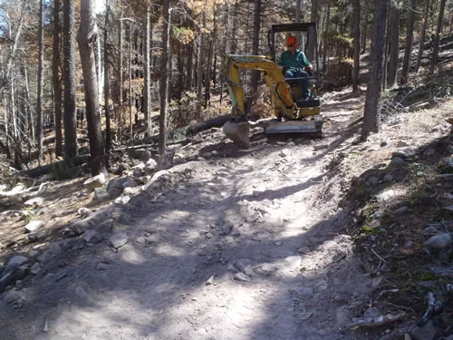 A mini excavator sits on a freshly benched trail on a steep forested slope, with cut logs and rock along the trail edge.