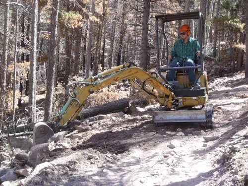 An operator in a hard hat uses a mini excavator to move a log off a freshly benched trail through standing burned timber.