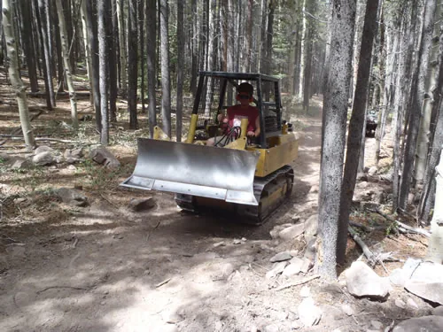 A yellow tracked trail dozer with an angled blade cuts new singletrack through a dense stand of aspen and conifer, operator seated under the roll cage.