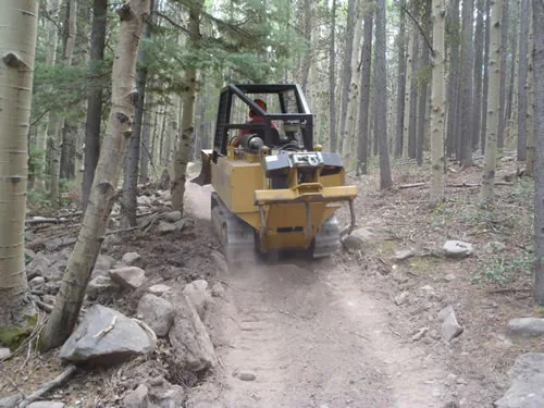 A tracked trail dozer climbs away up a freshly cut trail corridor through white-barked aspens, kicking up a plume of dust.