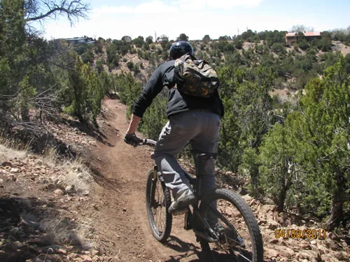 Mountain biker riding away from the camera down a narrow singletrack trail cut across a juniper-covered hillside