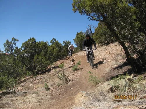 Two mountain bikers riding a narrow trail benched into a steep juniper-covered slope under a deep blue sky